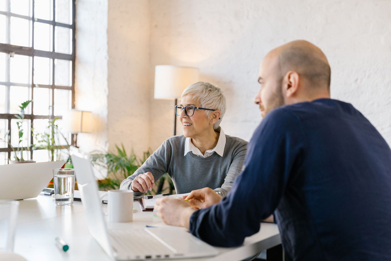 Eine erfahrene Frau und ein jüngerer Mann sitzen bei einem Beratungsgespräch an einem hellen Bürotisch mit Laptop in moderner