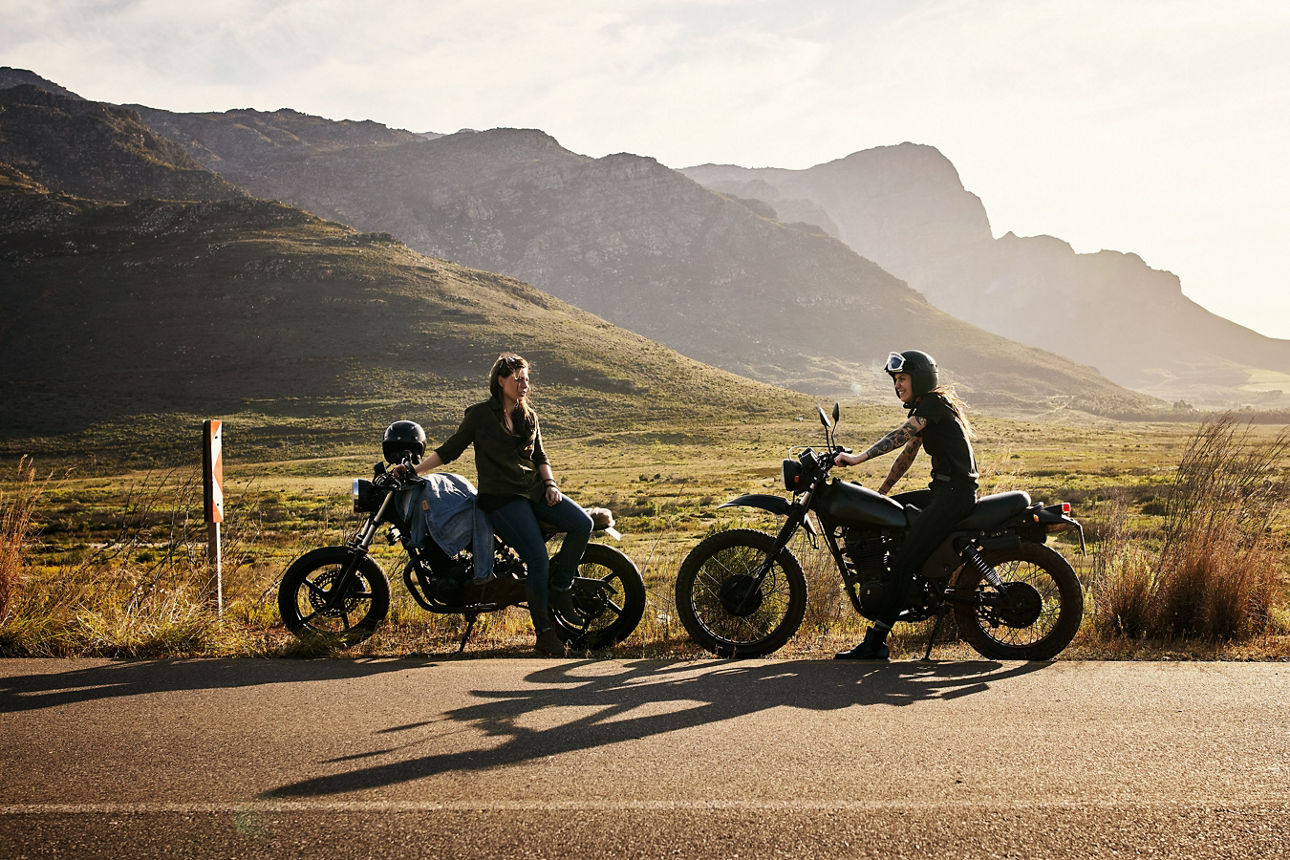 Zwei Motorradfahrerinnen stehen auf einer Landstraße hintereinander auf dem Seitenstreifen und machen eine kurze Pause. Im Hintergrund sieht man die Berge und es scheint die Sonne. Zwei Motorradfahrerinnen stehen auf einer Landstraße hintereinander auf dem Seitenstreifen und machen eine kurze Pause. Im Hintergrund sieht man die Berge und es scheint die Sonne.