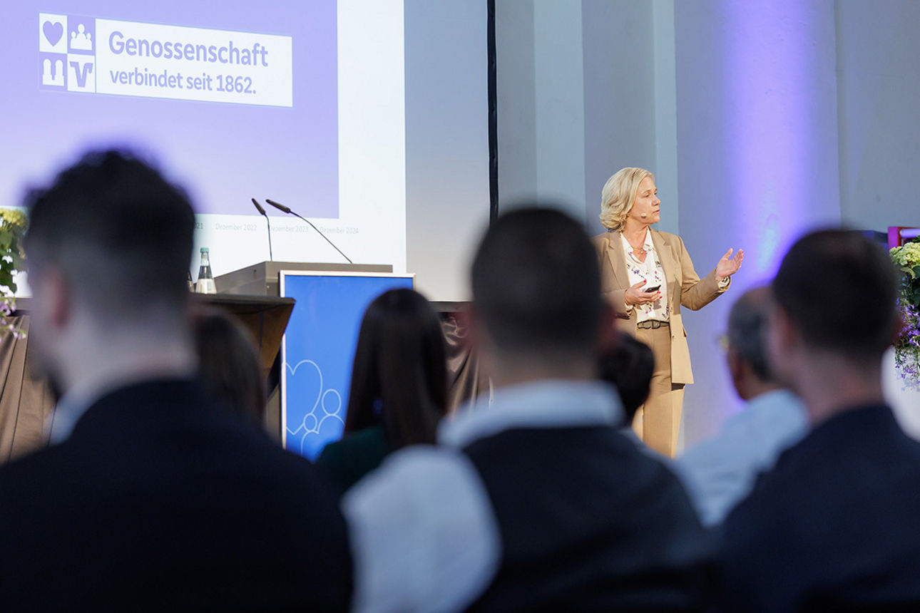 Eine Frau steht auf einem Podium und hält eine Rede, davor zahlreiche Gäste