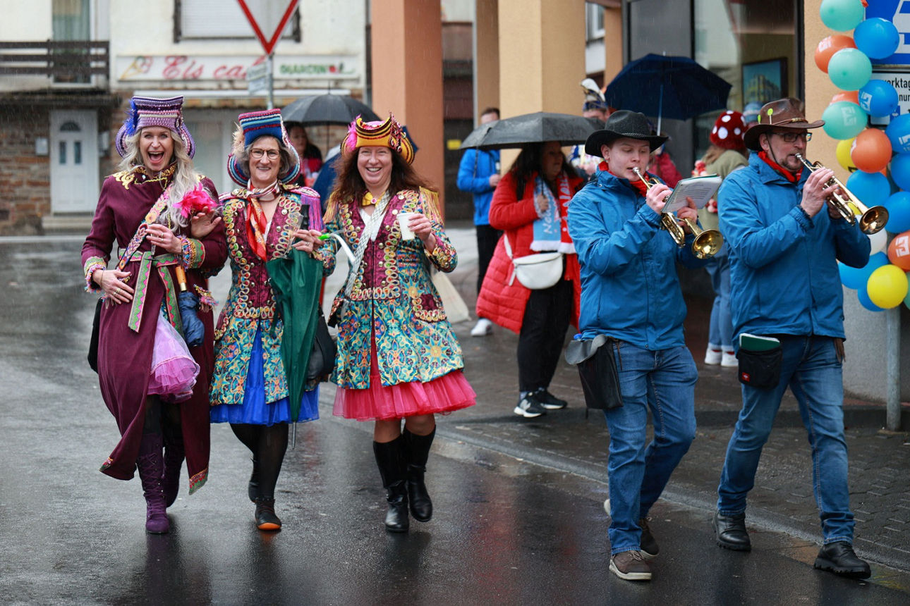 Drei Frauen in bunten Kostümen mit Hüten laufen Arm in Arm die Straße entlang