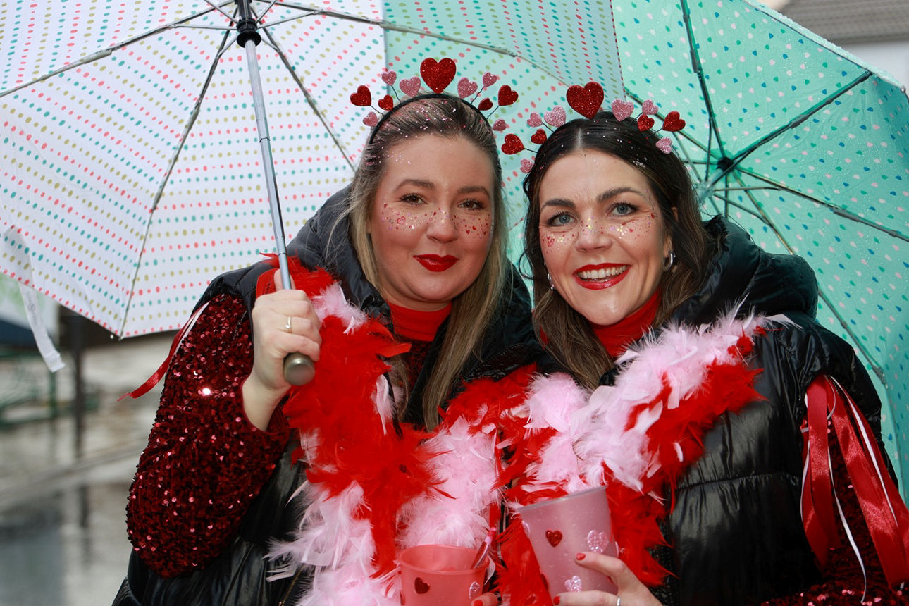 Zwei Frauen in rot-weißen Kostümen mit Federboa und Herzchen-Diademen unter bunten Regenschirmen