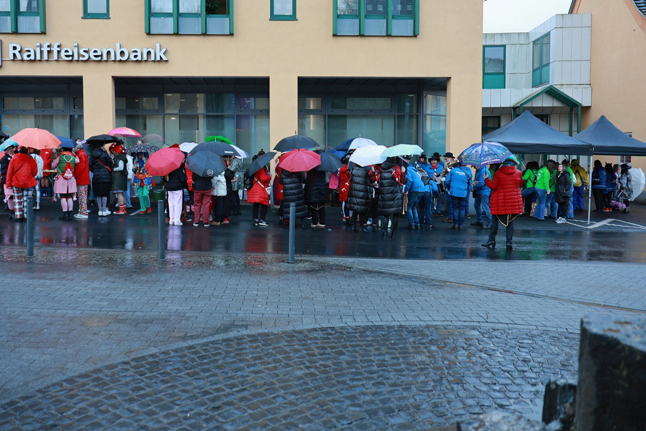 Die Narrenschar mit Regenschirmen hat sich vor dem Hauptgebäude der Bank in Neustadt versammelt