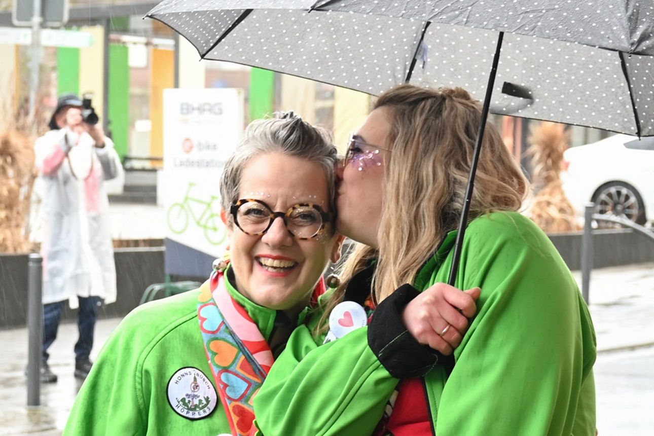Eine Frau mit blonden Haaren und grünem Pullover hält einen Regenschirm, während sie einer Frau einen Kuss auf die Stirn gibt