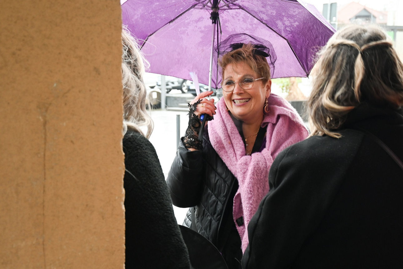 Eine Frau mit lila Schirm und rosa Schal steht mit einer Gruppe Frauen neben einem gelben Pfeiler
