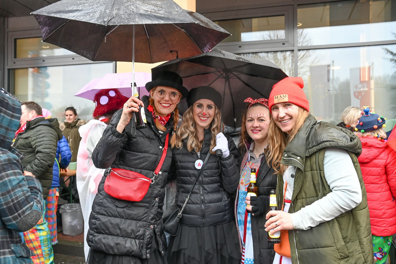 Vier Frauen in schwarzen Harlekin- und bunten Clowns-Kostümen stehen unter schwarzen Regenschirmen und lächeln in die Kamera