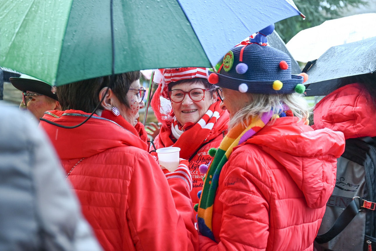 Drei Frauen stehen unter einem grünen Schirm zusammen und unterhalten sich. Alle tragen bunte Kostüme und rote Jacken.