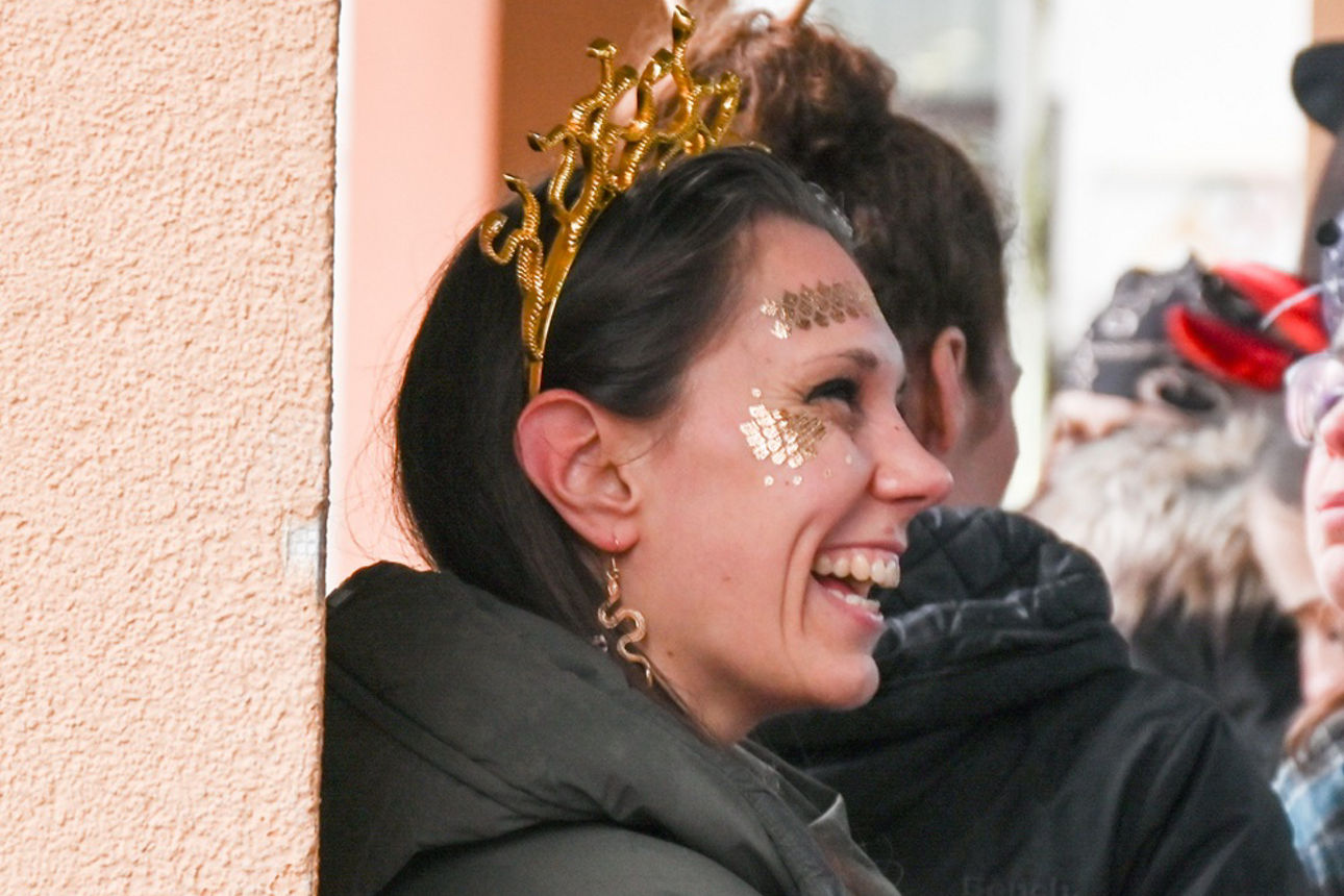 Eine junge Frau im goldenen Medusa-Kostüm lehnt an einem gelblichen Pfeiler und lacht fröhlich; Profilaufnahme