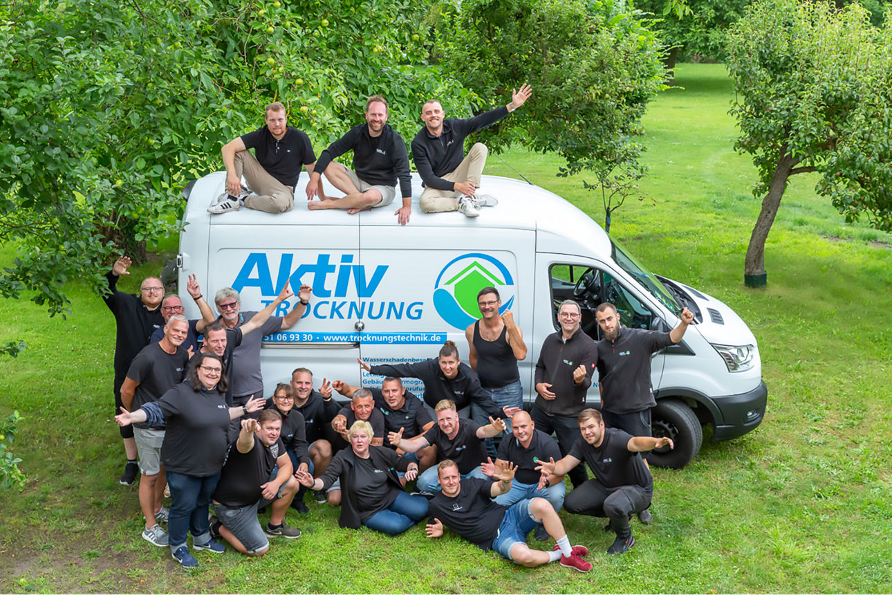 Team der "Aktiv Trocknung" posiert lachend vor ihrem Firmenwagen in einer grünen Parkanlage.