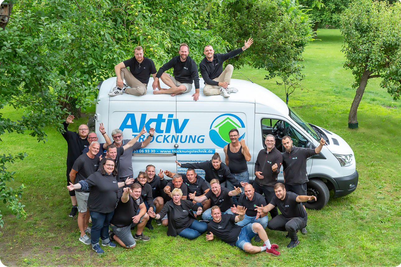 Team der "Aktiv Trocknung" posiert lachend vor ihrem Firmenwagen in einer grünen Parkanlage.