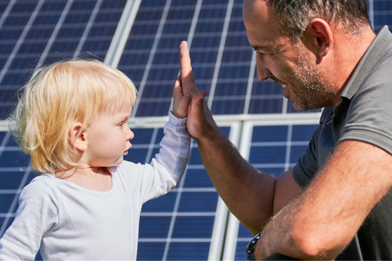 Erwachsene Person und Kleinkind geben sich vor einer großen Solaranlage ein High‑Five im Grünen.
