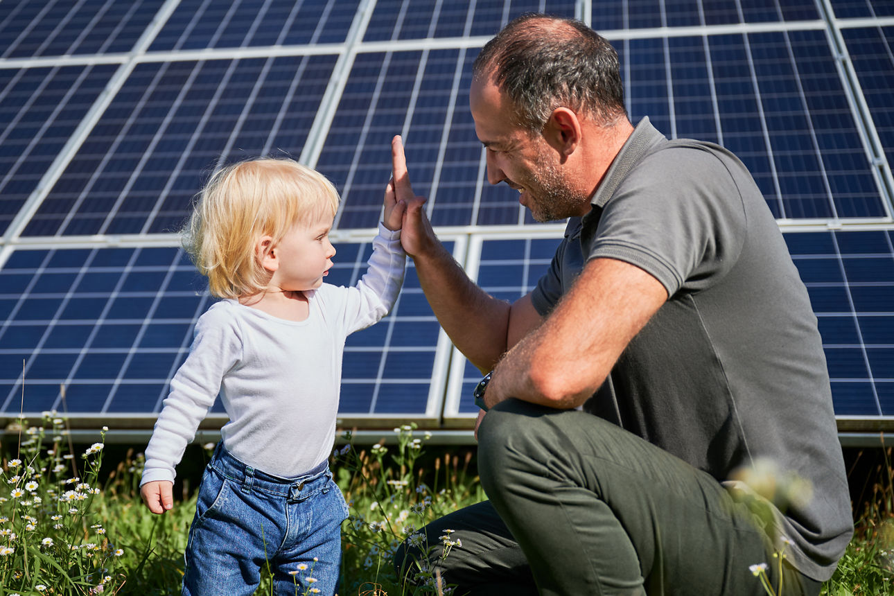 Erwachsene Person und Kleinkind geben sich vor einer großen Solaranlage ein High‑Five im Grünen.