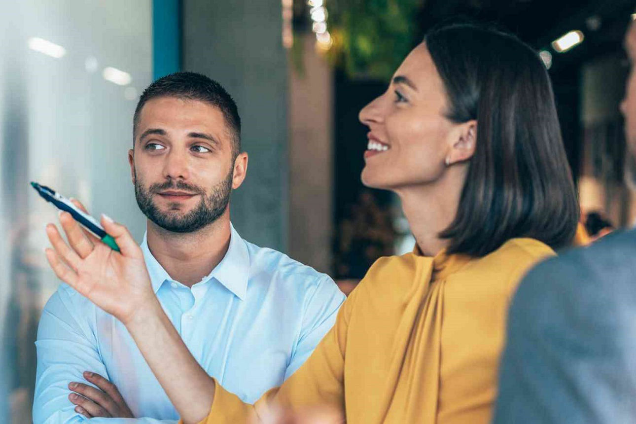 Arbeitskollegen stehen vor einer Tafel. Eine Frau hält einen Stift in der Hand und deutet auf die Tafel.