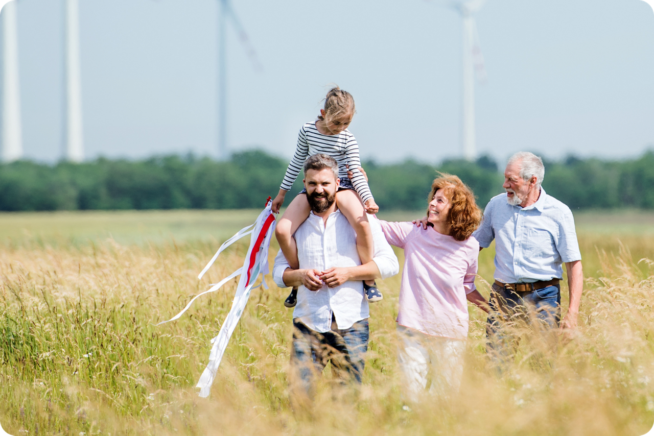 Drei Personen laufen durch eine Wiese, auf der Schulter eines Mannes sitzt ein Kind, im Hintergrund sind Windräder zu sehen