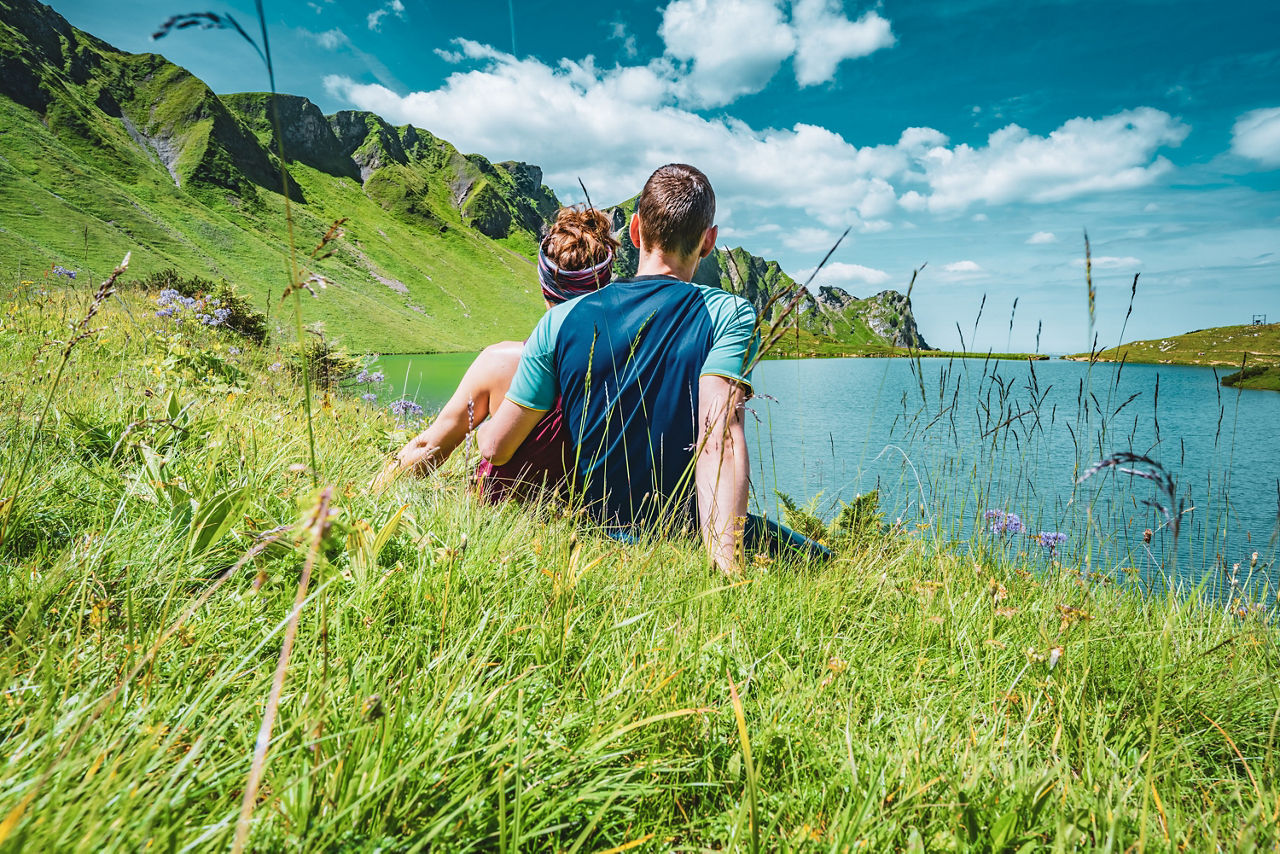Ein Paar sitzt auf einer Wiese am Berg. Sie blicken gemeinsam in Richtung See.