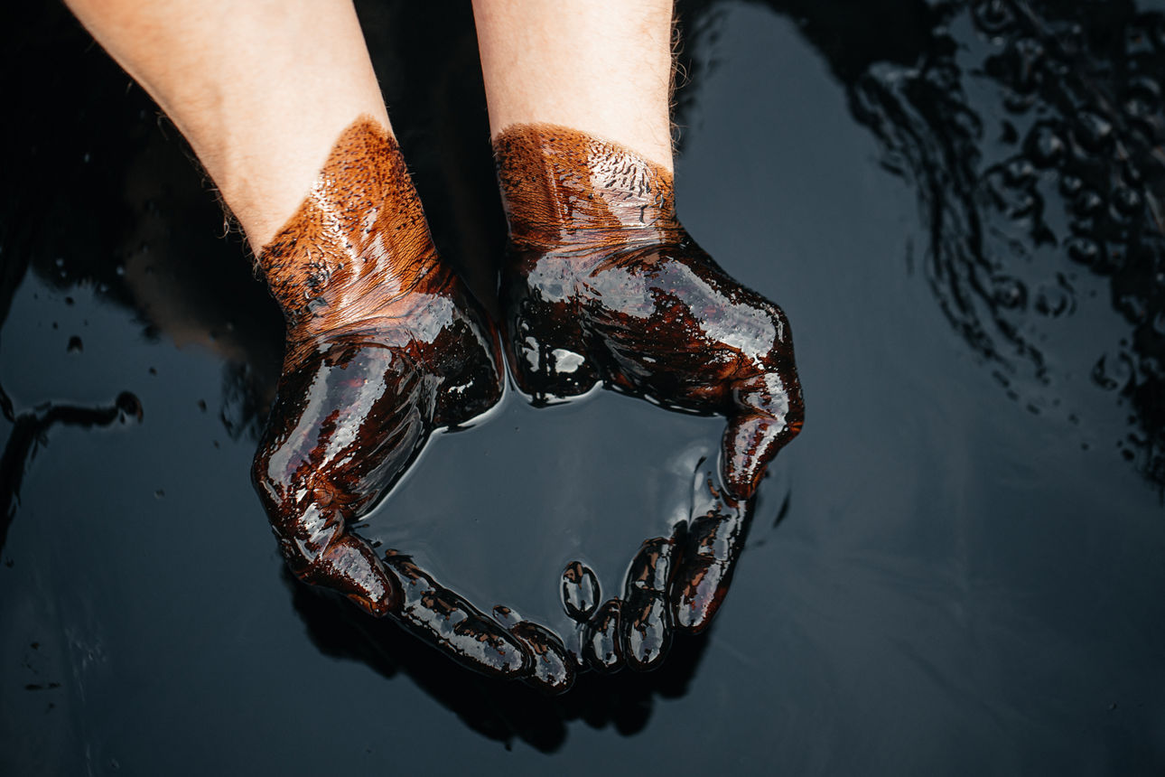 Zwei Hände, mit schwarzem Öl verschmutzt, formen eine Schale und halten eine kleine Menge Flüssigkeit.