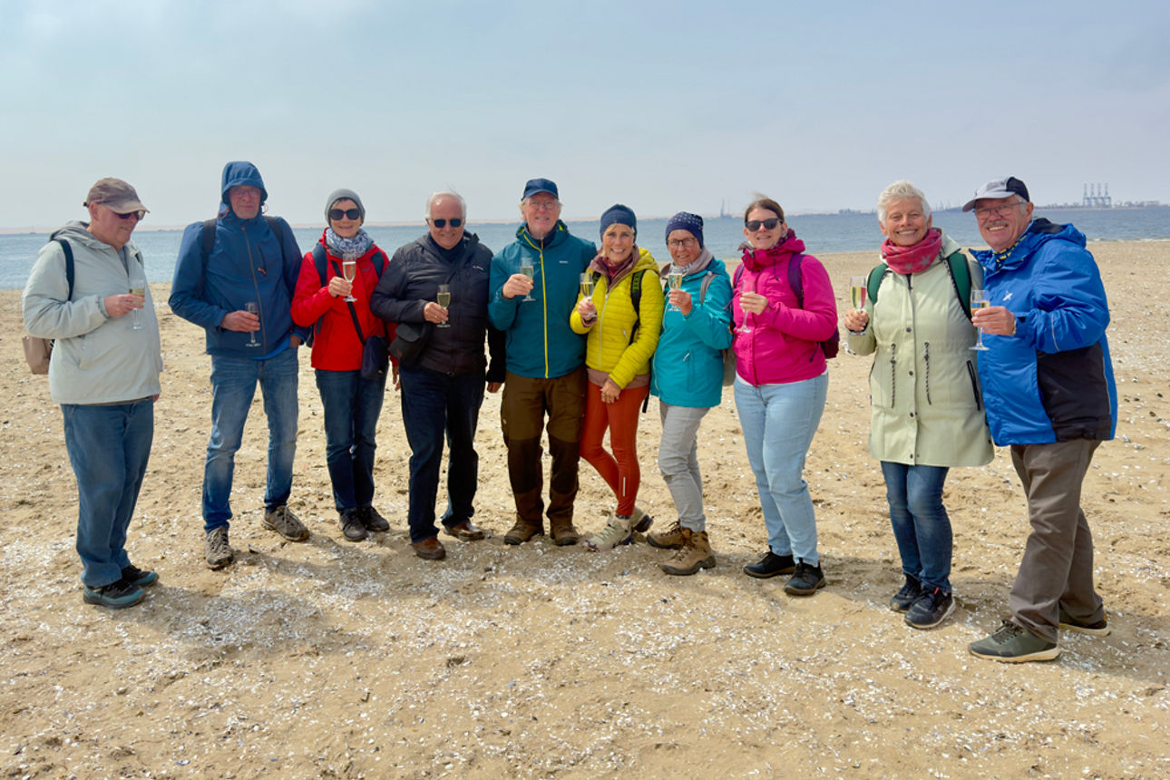 Eine Gruppe von zehn Touristen in farbenfroher Kleidung stößt am Strand mit Champagnergläsern an.
