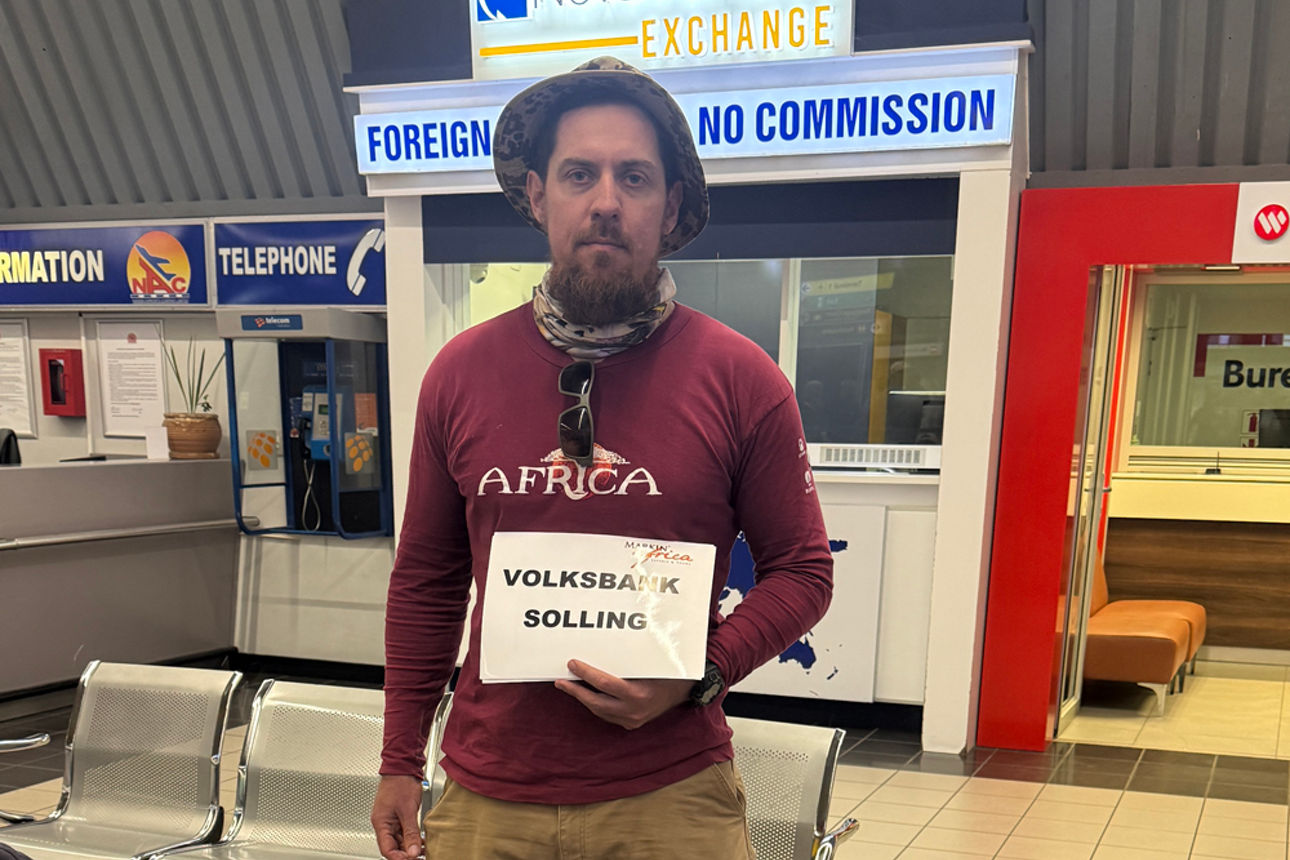 Mann mit Schild "Volksbank Solling" am Flughafen, davor Gepäck.