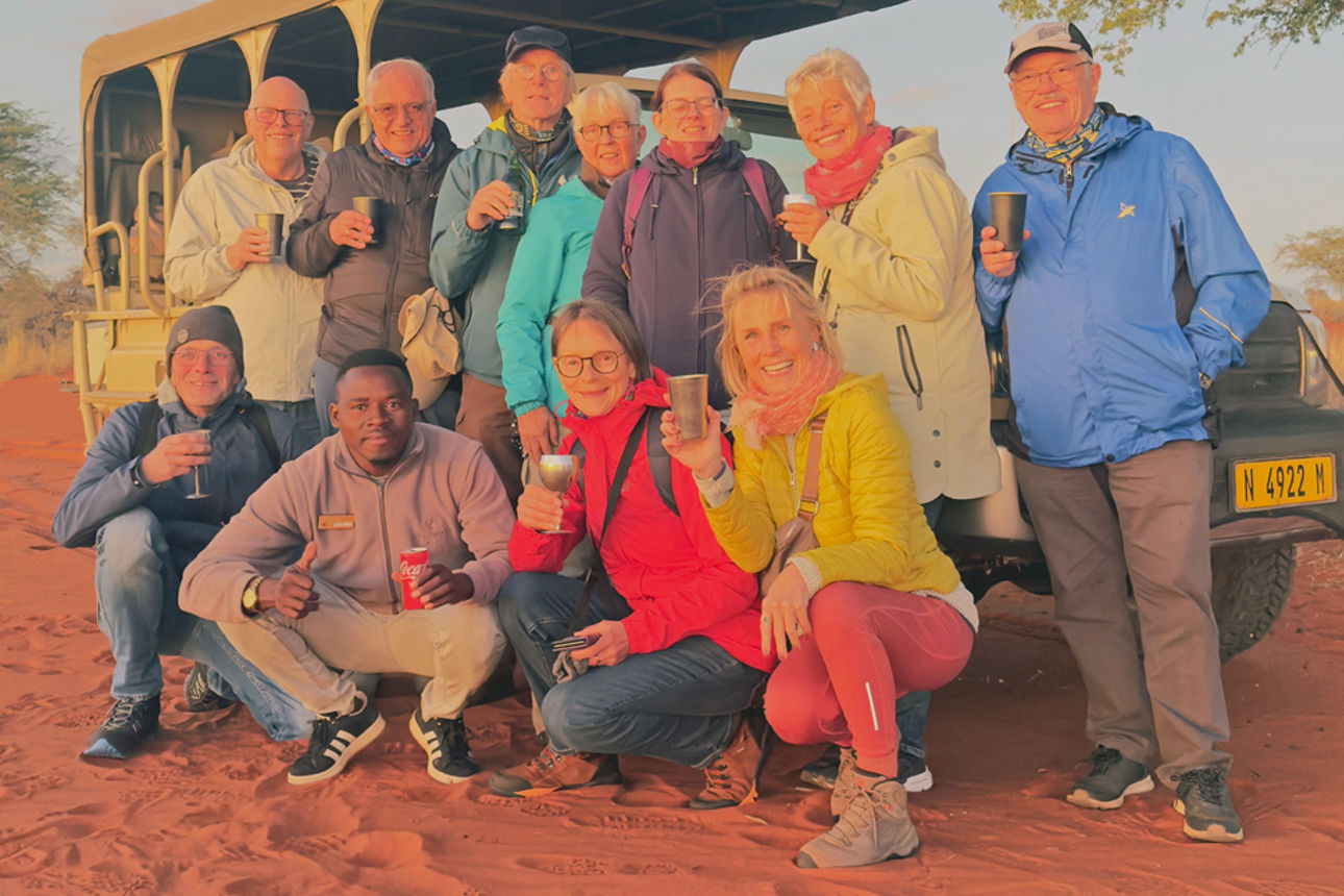 Reisegruppe stößt vor einem Safarifahrzeug auf rotem Sand an.