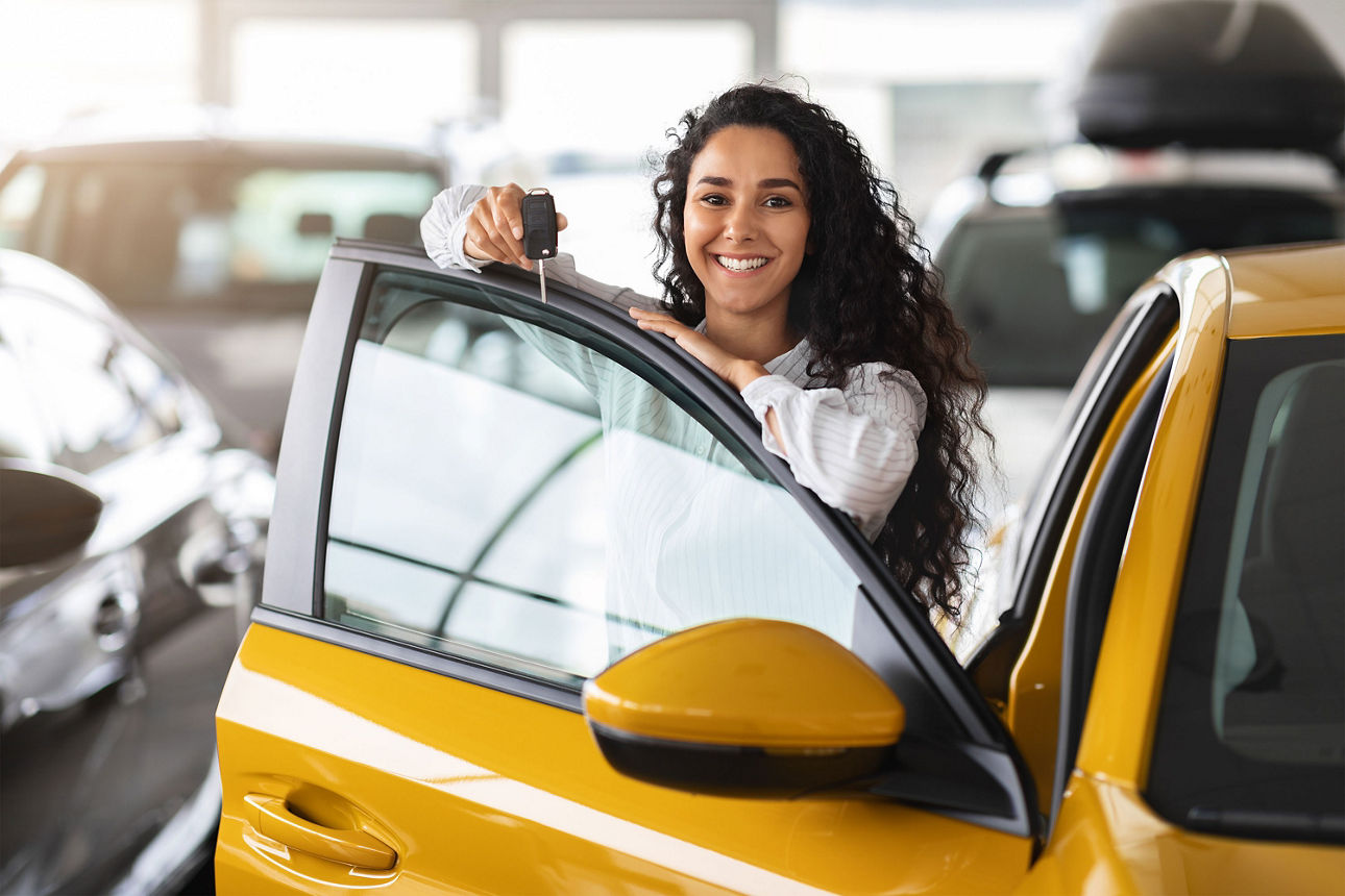 Eine fröhliche brünette Frau lehnt sich auf eine Autotür und hält einen Autoschlüssel in der Hand