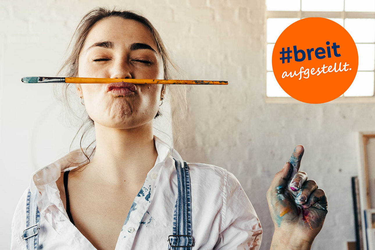 Eine junge Frau steht mit Farbflecken auf der Kleidung und einem Pinsel zwischen Mund und Nase geklemmt in einem Atelier