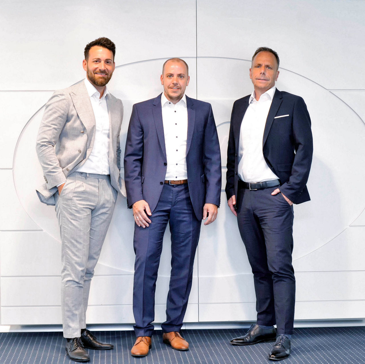 3 Männer in Anzügen stehen vor einer Weißen Wand