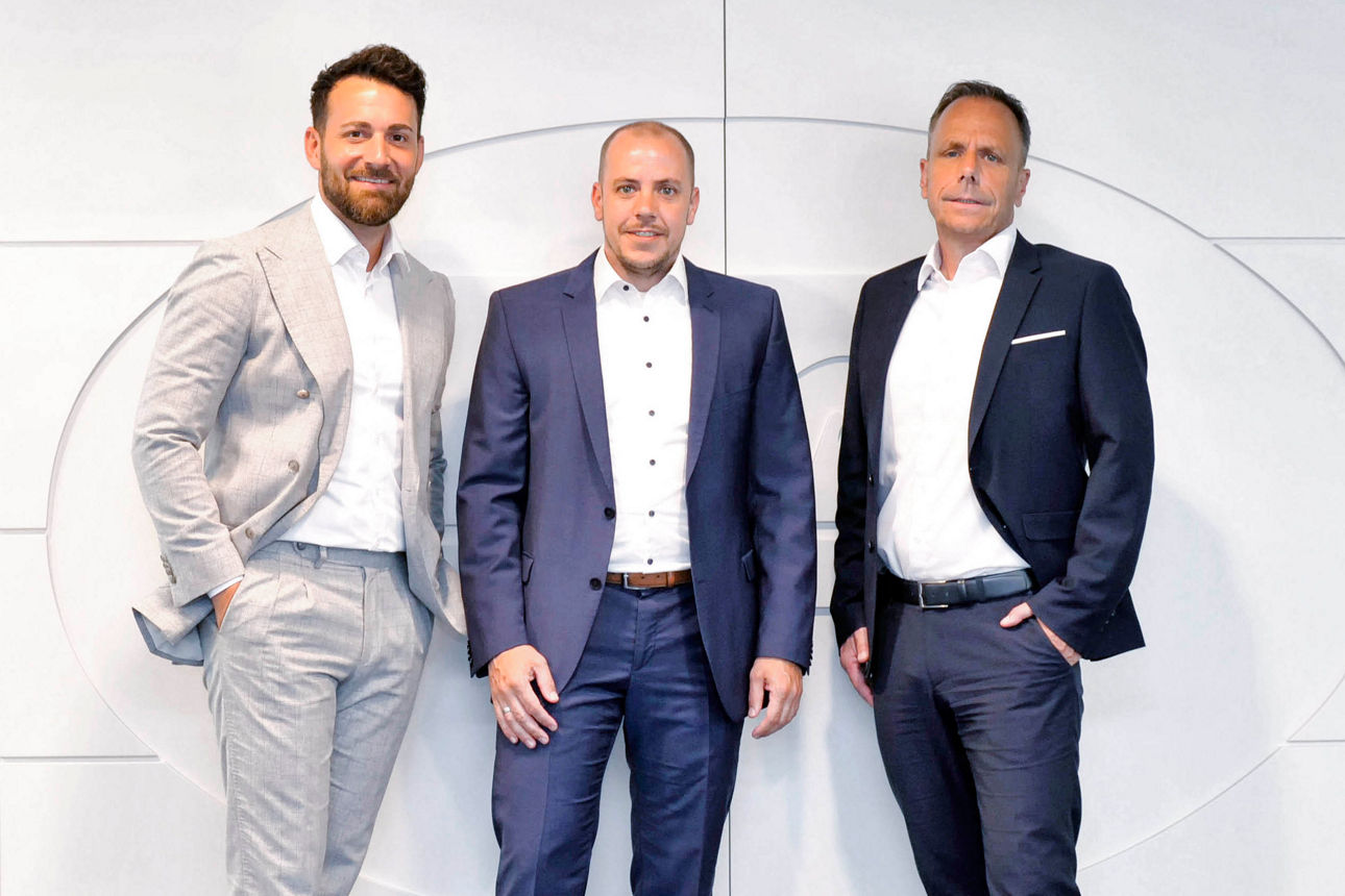 Drei Herren im Anzug vor einem hellen Hintergrund. 