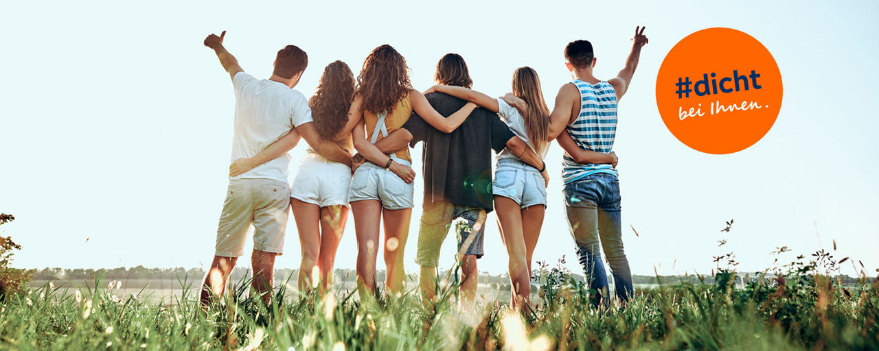 Junge Leute von hinten stehen auf einer Wiese dicht beieinander und freuen sich