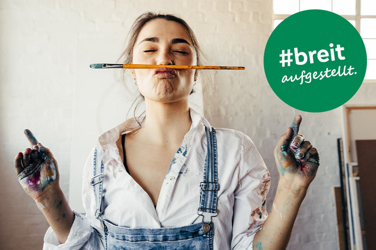 Eine junge Frau steht mit Farbflecken auf der Kleidung und einem Pinsel zwischen Mund und Nase geklemmt in einem Atelier