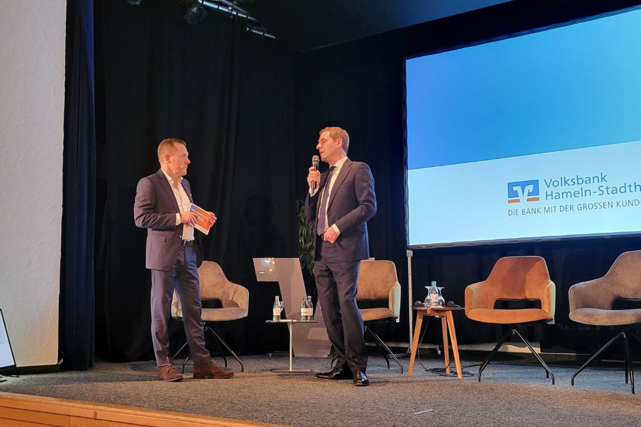 Auf einer Bühne führen zwei Personen in Anzügen ein Gespräch mit Mikrofon.