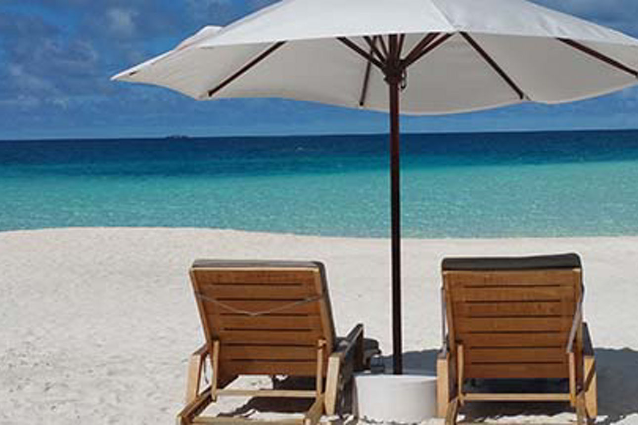 Zwei Liegestühle unter einem großen Sonnenschirm, im Hintergrund weicher Sand und ein strahlend blauer Himmel. 