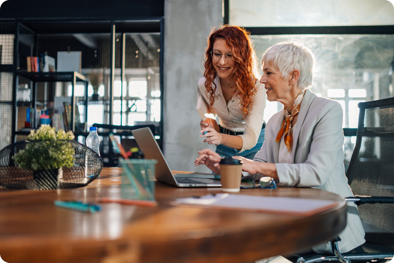 Zwei Frauen stehen und sitzen an einem Tisch vor einem Laptop und diskutieren