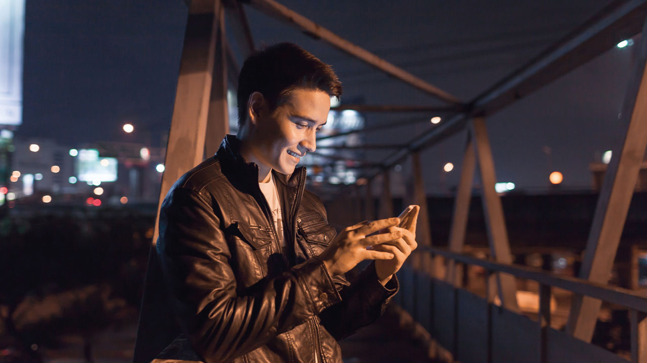 Junger Mann lehnt bei Nacht auf einer Metallbrücke am Geländer und schaut auf sein Handy. Skyline einer Stadt im Hintergrund