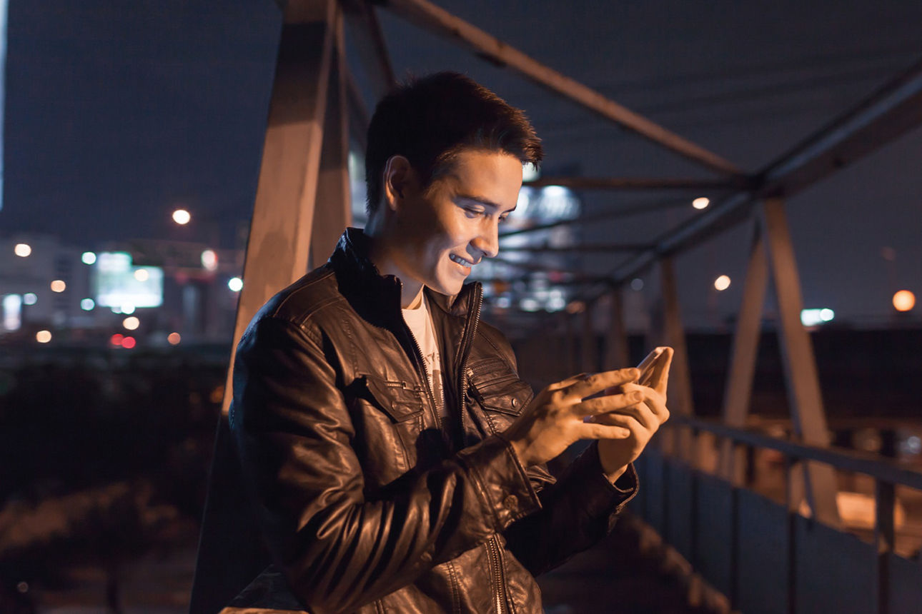 Junger Mann lehnt bei Nacht auf einer Metallbrücke am Geländer und schaut auf sein Handy. Skyline einer Stadt im Hintergrund