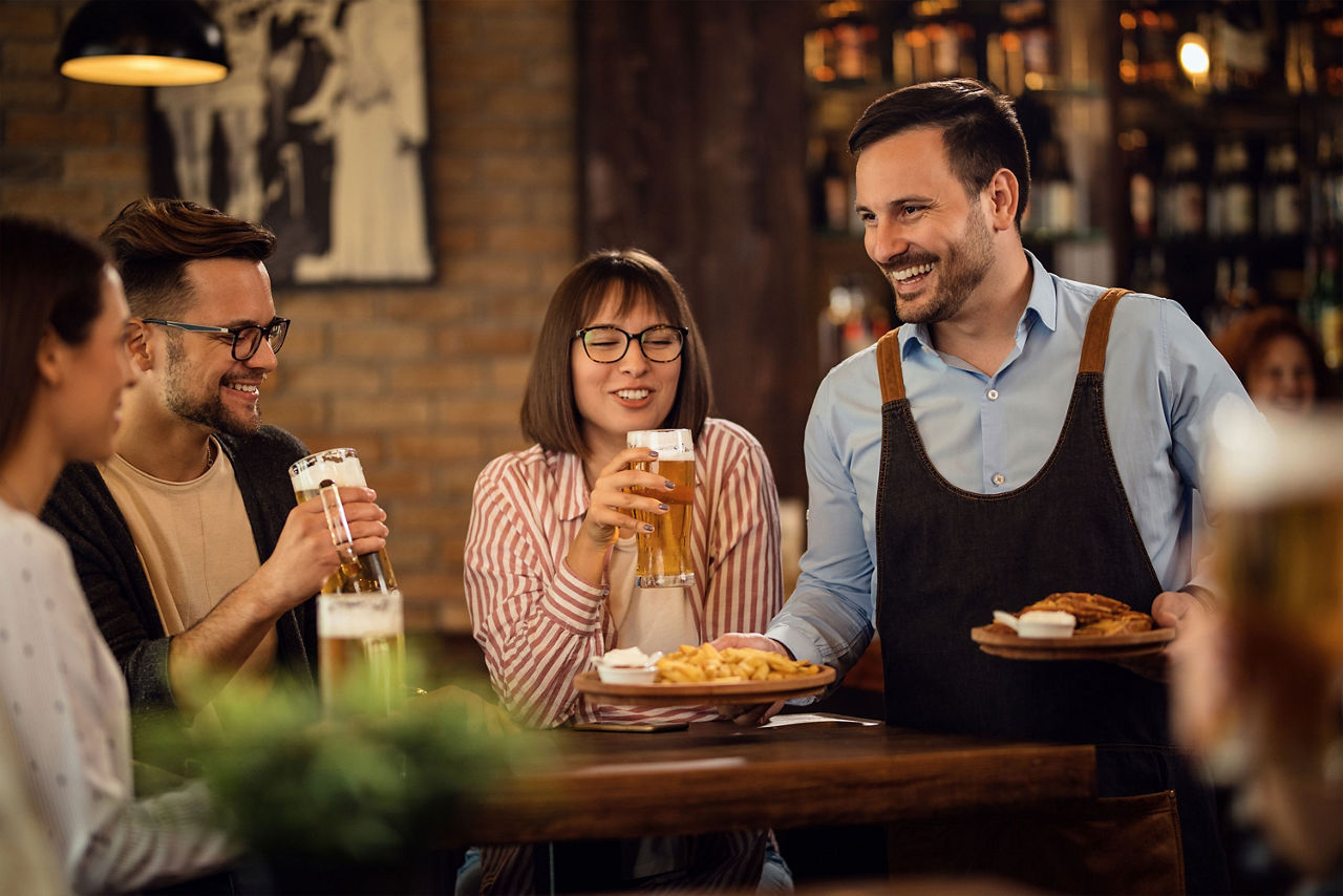 Ein fröhlicher Kellner bringt einer Gruppe von Leuten, die in einer Kneipe Bier trinken, das Essen.