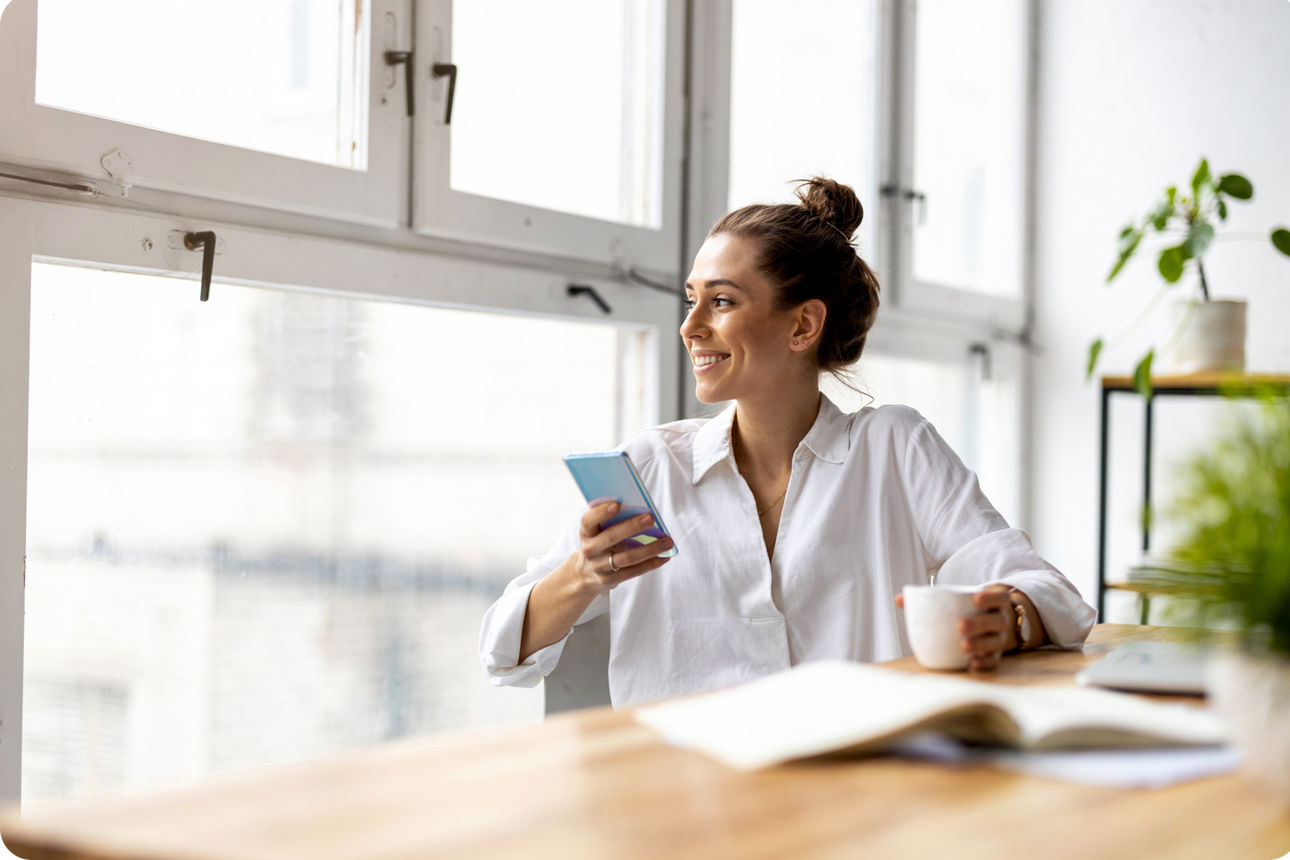 Person sitzt am Tisch, hält ein Smartphone und eine Tasse vor einem großen Fenster.