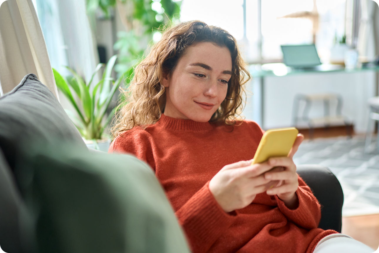 Person sitzt auf einem Sofa und nutzt ein Smartphone in einem hellen Wohnzimmer.
