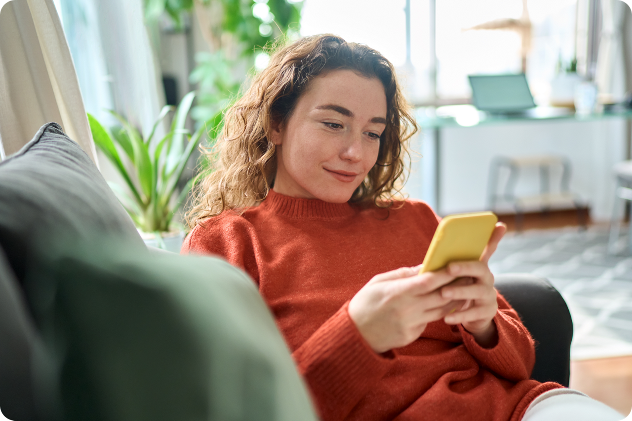 Person sitzt auf einem Sofa und nutzt ein Smartphone in einem hellen Wohnzimmer.