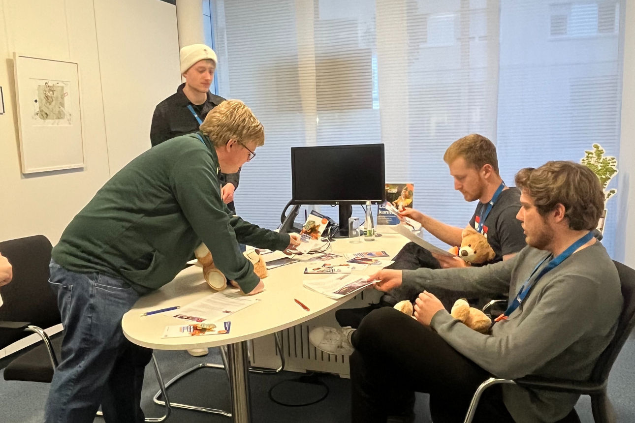 Gruppe sitzt am Tisch und betrachtet Infomaterial sowie Stofftiere in einem Büro.