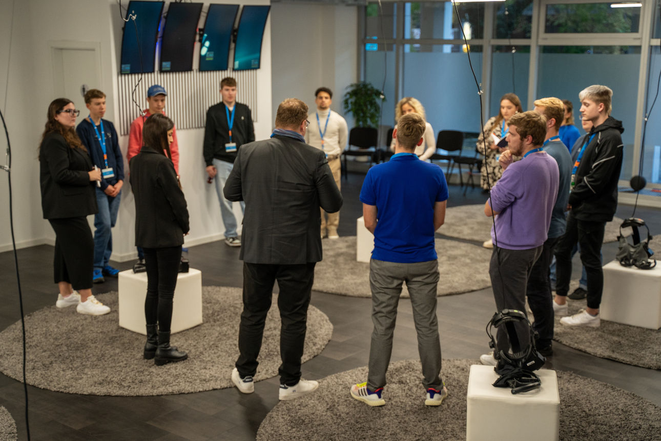 Teilnehmende stehen im Kreis in einem Technikraum und hören einer Präsentation zu.