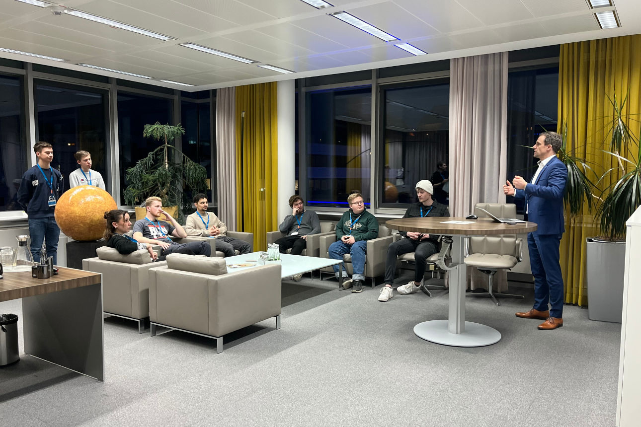 Gruppe sitzt in einer Lounge, während eine Person vorne einen Vortrag hält.