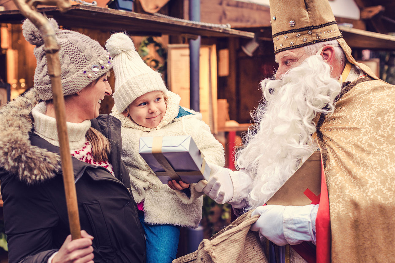Frau mit Kind auf dem Arm bekommt auf einem Weihnachtsmarkt ein Geschenk von einem Nikolaus.