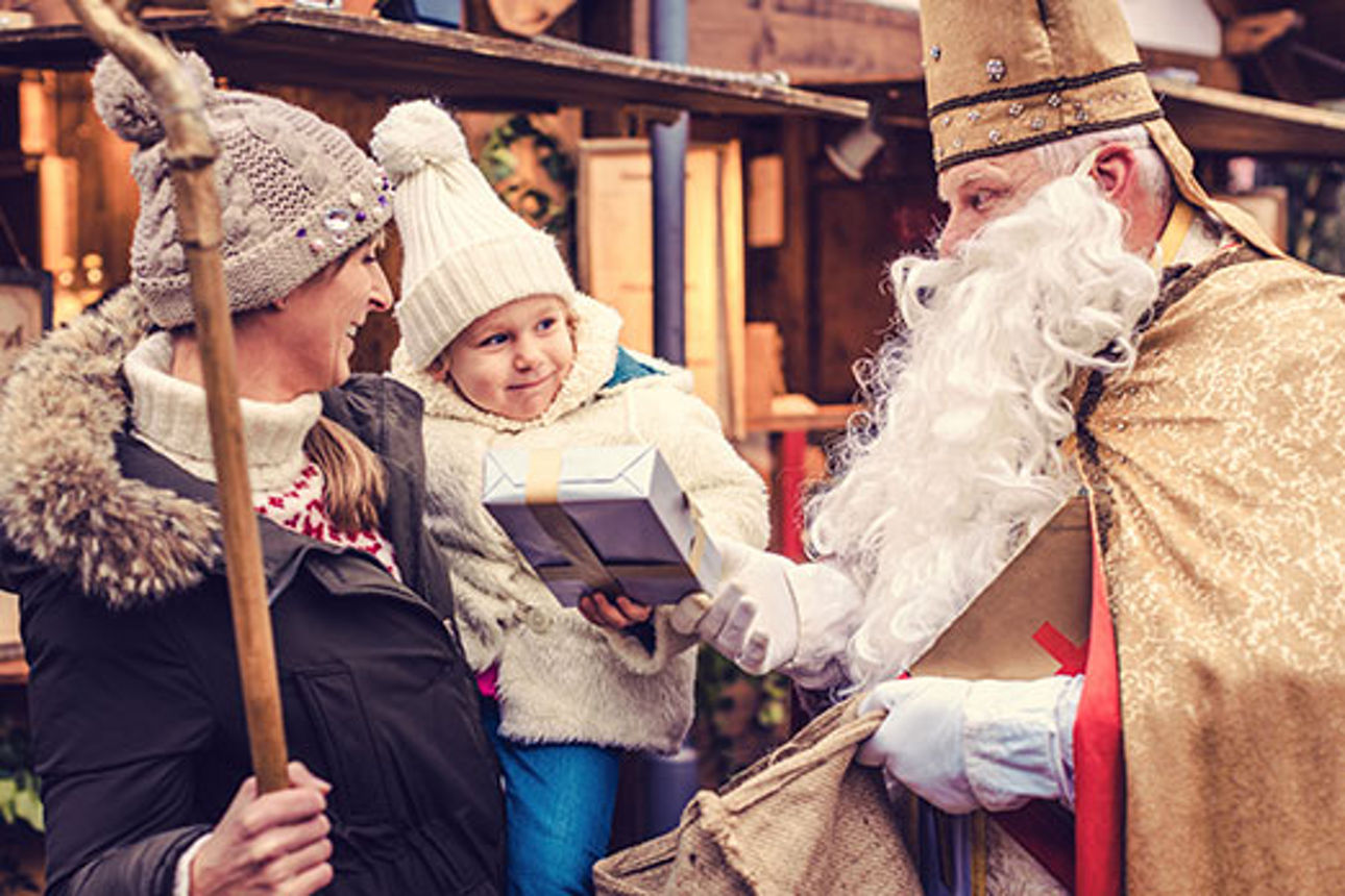 Eine weihnachtlich gekleidete Person übergibt einem Kind ein Geschenk vor festlich geschmückten Marktständen.