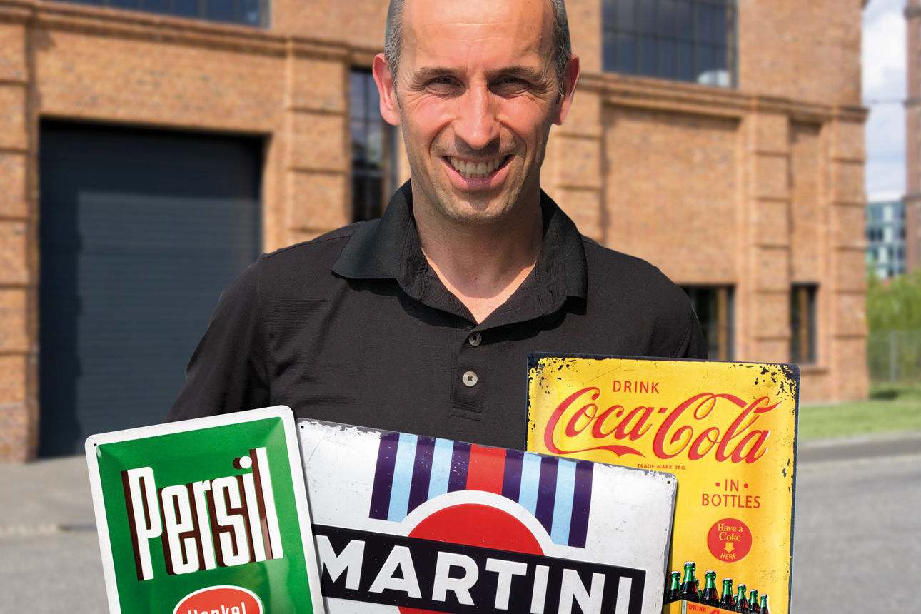 Ein Mann steht vor einem alten Haus auf der Straße und hält 3 nostalgische Metallschilder hoch