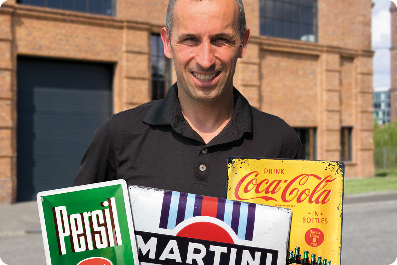 Ein Mann steht vor einem alten Haus auf der Straße und hält 3 nostalgische Metallschilder hoch