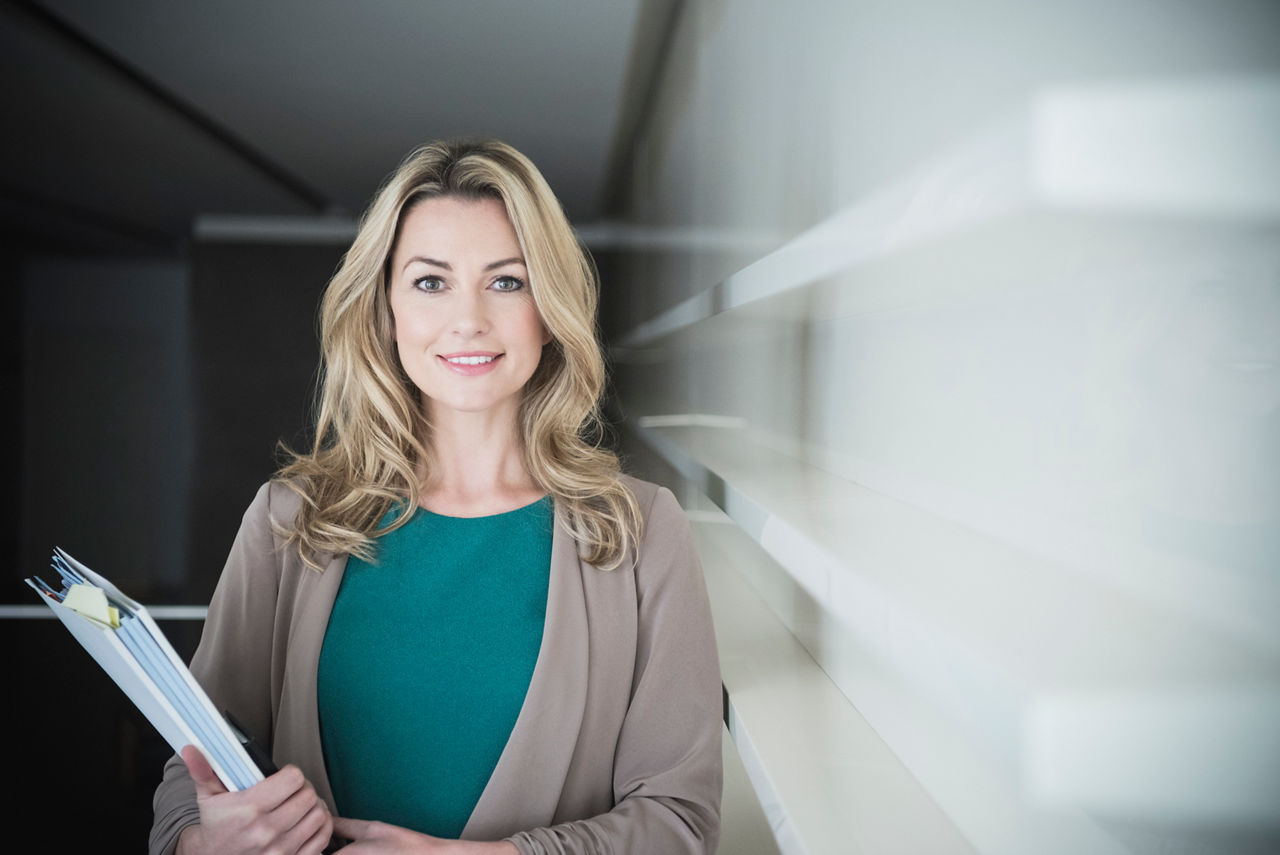 Weibliche Büroangestellte mit langen blonden Haaren schaut in die Kamera und lächelt. Frau hält Dokumente in einem modernen Büro in den Händen
