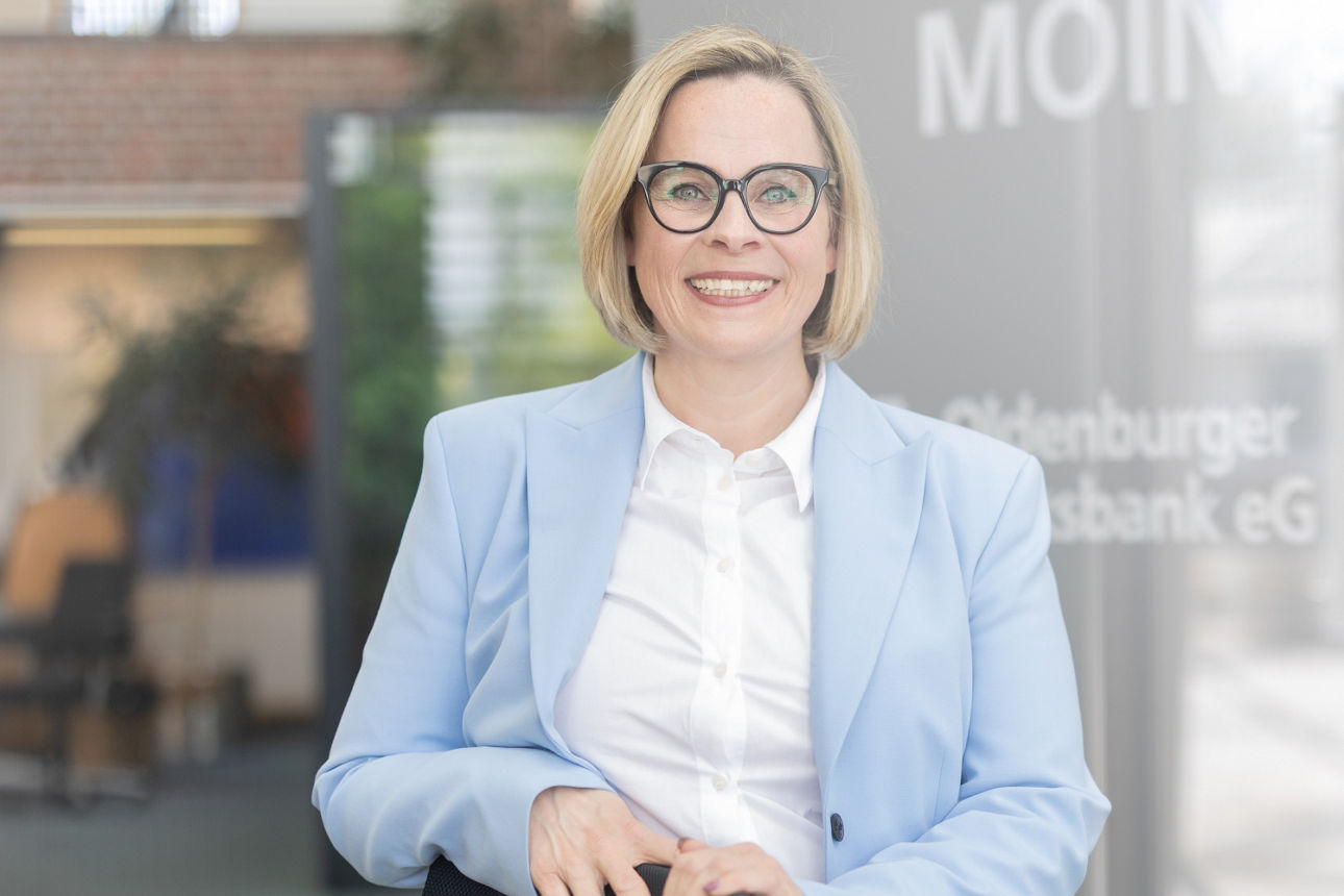 Eine Frau in hellem Blazer steht lächelnd in einem Gebäude