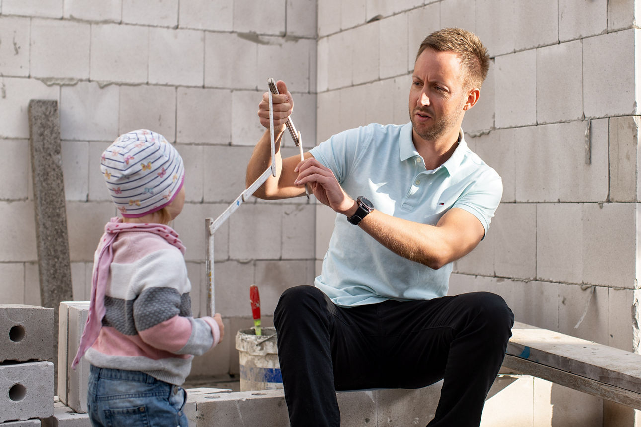 Mann sitzt mit Kind vor dem Rohbau eines Hauses