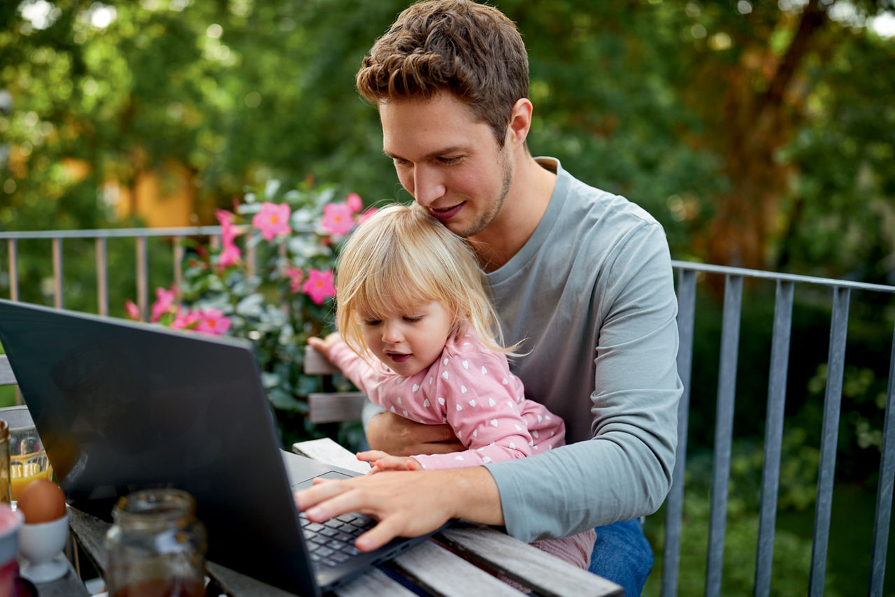 Vater sitzt mit Tochter auf dem Schoß auf dem Balkon und arbeitet am Laptop