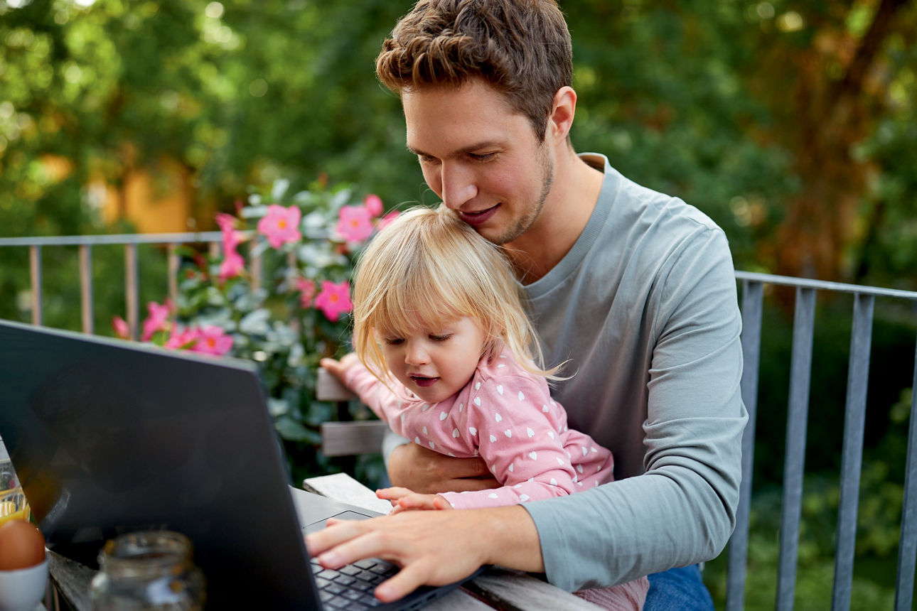 Vater sitzt mit Tochter auf dem Schoß auf dem Balkon und arbeitet am Laptop