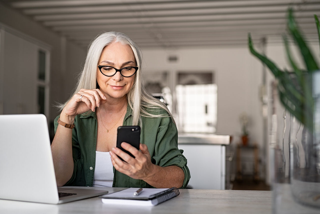 Frau vor einem Laptop, mit einem Smartphone in der Hand